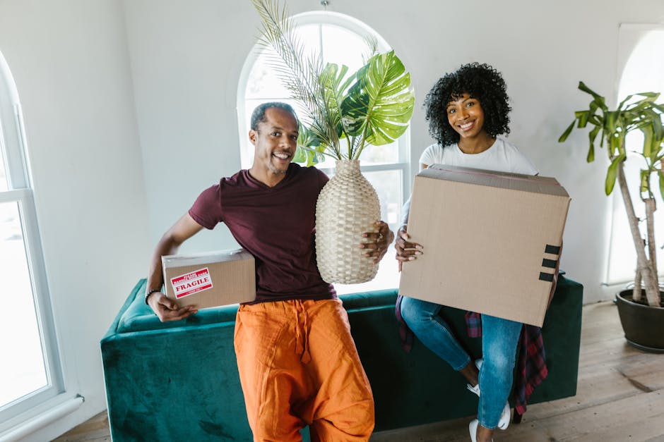 A man and woman are engaged in a home relocation process outside a building, facing each other closely with smiling expressions. The man, with short curly dark hair and wearing a maroon t-shirt, is ho