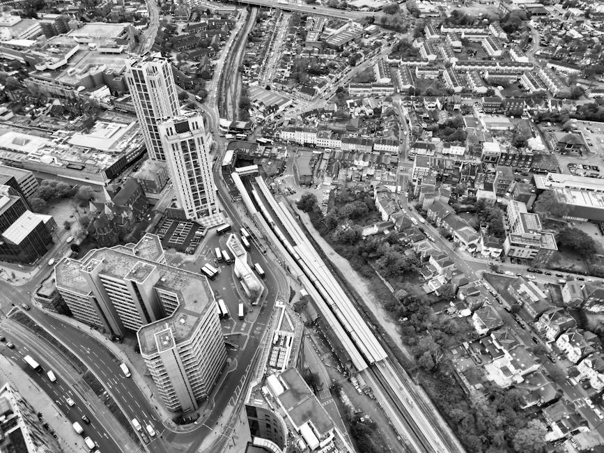 Aerial black-and-white photograph of an urban area featuring a cluster of high-rise buildings, smaller commercial and residential structures, and a network of roads and railway tracks. A train station