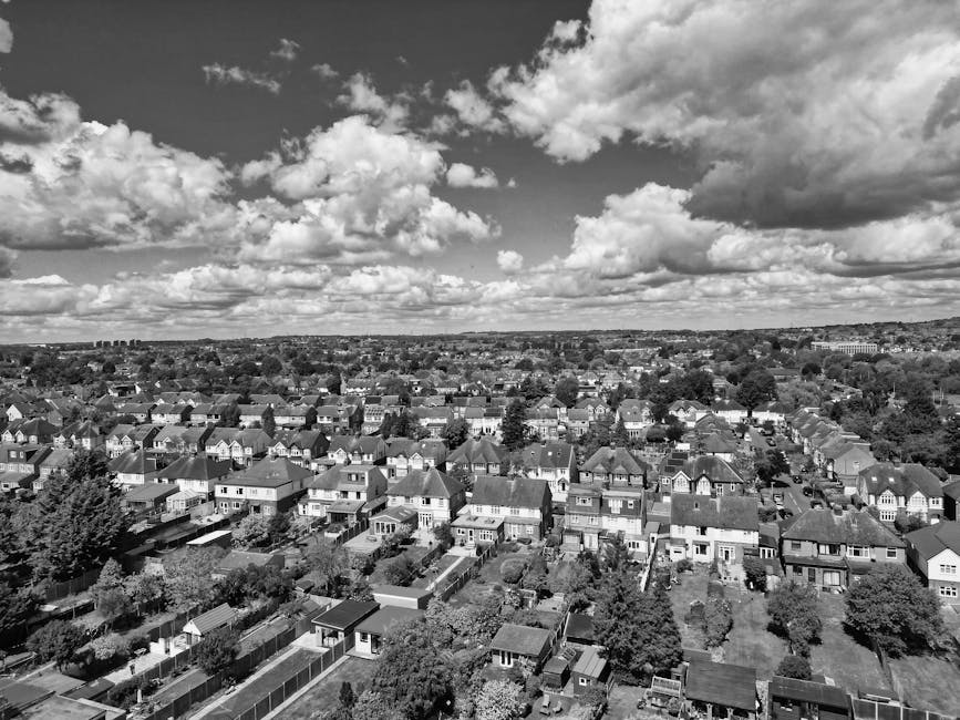 Aerial black-and-white photograph of an urban area featuring a cluster of high-rise buildings, smaller commercial and residential structures, and a network of roads and railway tracks. A train station