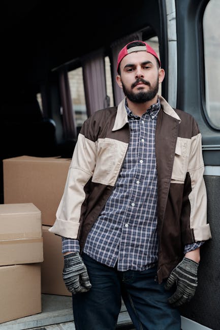 A man with a beard and wearing a red cap, checked shirt, and a beige and brown jacket stands inside the open back of a van used for house removals. Behind him are stacked cardboard boxes and packing materials, including wrapped furniture. The van's interior shows a spacious cargo area with additional boxes nearby, ready for loading or unloading during a home relocation. The scene is outdoors, with natural lighting illuminating the area, and the man appears to be part of a professional moving process associated with Dagenham Man and Van, providing furniture transport and packing services on the Becontree Estate Dagenham area.