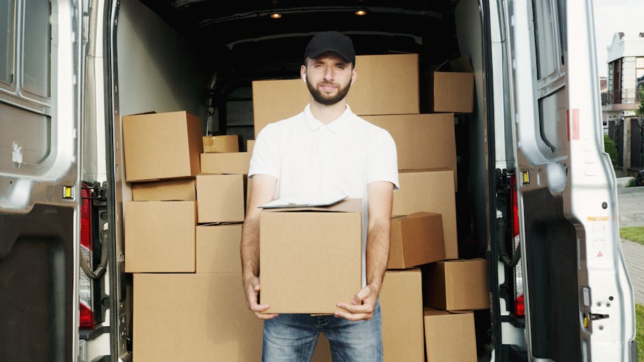 A man wearing a black cap, white polo shirt, and jeans stands inside the open rear door of a white van loaded with various cardboard boxes, some stacked on top of each other and others arranged around him. He is holding a medium-sized box with both hands and has a neutral expression. The van's interior shows additional boxes and packing materials, indicating a home relocation or furniture transport process. Outside, part of a paved driveway and surrounding residential buildings are visible, with natural daylight illuminating the scene. The setup suggests professional removals services, such as those offered by Dagenham Man and Van, involved in packing and moving tasks typical of house removals within the Becontree Estate area.