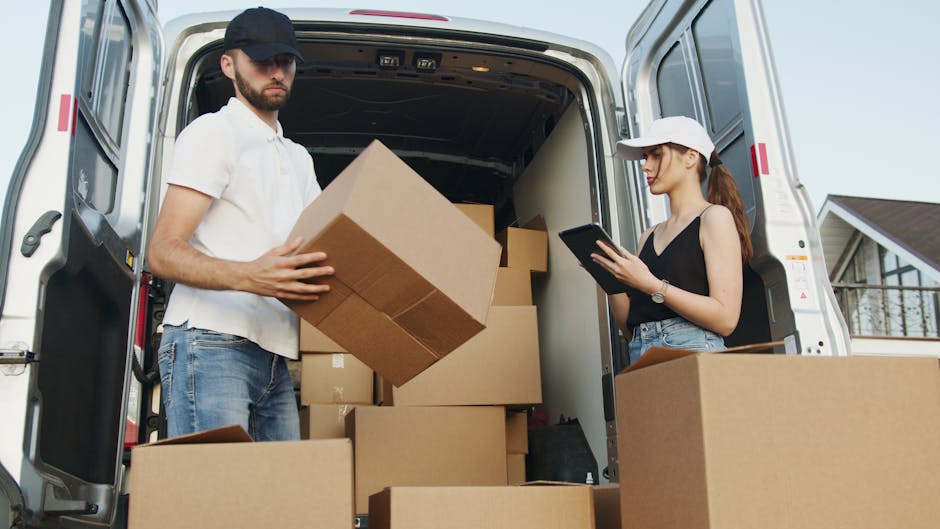 A man with a beard and wearing a red cap, checked shirt, and a beige and brown jacket stands inside the open back of a van used for house removals. Behind him are stacked cardboard boxes and packing materials, including wrapped furniture. The van's interior shows a spacious cargo area with additional boxes nearby, ready for loading or unloading during a home relocation. The scene is outdoors, with natural lighting illuminating the area, and the man appears to be part of a professional moving process associated with Dagenham Man and Van, providing furniture transport and packing services on the Becontree Estate Dagenham area.