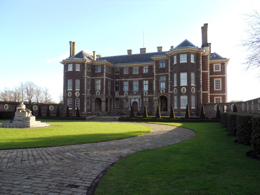 A large, historic brick mansion with multiple stories, many windows, and decorative stonework, situated behind a well-maintained lawn and a curved brick pathway. The building has a peaked roof with chimneys, and a stone statue is visible on a pedestal to the left side of the lawn. The scene is lit by natural daylight, with clear skies and some trees visible in the background. This setting reflects a substantial property that may be involved in home relocations or estate removals, with the exterior prepared for logistics such as packing, furniture transport, and loading onto vehicles, aligning with services offered by Dagenham Man and Van for removals near Valence House Museum Dagenham.