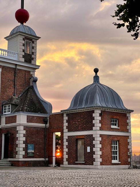 Photograph of Valence House Museum in Dagenham during sunset, showcasing its historic brick facade with decorative white stone accents around the windows and edges. The building features a distinctive domed roof with a metallic finish, a small tower with a red weather vane or finial, and a surrounding stone-paved area. The sky displays a mix of orange, yellow, and purple hues, indicating late evening light. In the foreground, part of the paved courtyard and a black metal fence are visible, with some foliage and leaves from nearby trees partially framing the image. The scene highlights the architectural detail of the museum, emphasizing its historical character and the atmospheric evening setting, with no visible moving activity or furniture included.