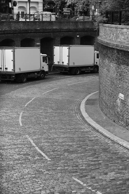 A black and white photograph showing two medium-sized box trucks parked on a cobblestone driveway adjacent to a brick wall and a curved street. The trucks are positioned side by side, with the rear of each vehicle facing away from the camera, indicating they are in the process of loading or unloading. Behind the trucks, there is a stone wall with arched openings, possibly leading to an underground or basement exit, and above this wall, a balcony or terrace with railings and some trees visible in the background. The scene appears to be part of a home relocation or furniture transport operation, with the trucks likely used for a local removals service by Dagenham Man and Van, associated with the page for same-day removals in Dagenham Dock. The environment is well-lit, highlighting the movement of goods typical in packing and moving logistics.