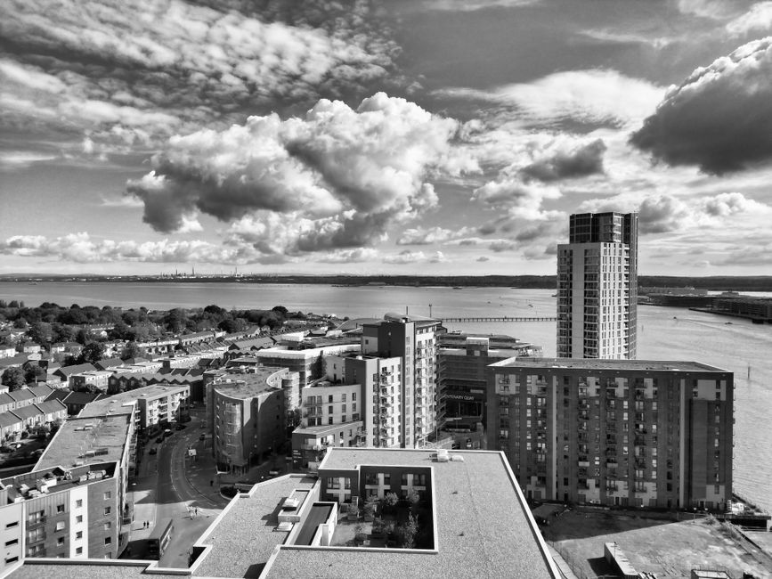 A black-and-white aerial view of a waterfront urban area showing several modern multi-storey buildings, some with balconies and rooftop terraces, situated along a curved street. In the foreground, a flat roof with a small enclosed garden or courtyard can be seen. To the right, there is a tall, narrow residential building next to a river or large body of water, with a bridge spanning across it in the distance. The scene features dramatic cloud formations in the sky and is illuminated by natural daylight, highlighting the structural details of the buildings. This setting illustrates the type of environment where house removals and furniture transport might occur, aligning with services for home relocation or moving logistics offered by Dagenham Man and Van.